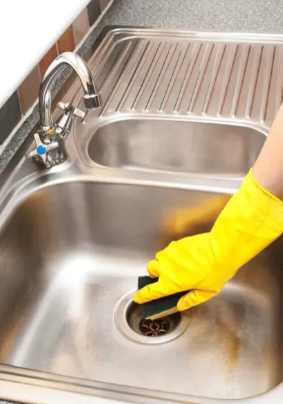 A person wearing a bright yellow rubber glove uses a scrubbing sponge to clean the drain area of a double-basin stainless steel kitchen sink.